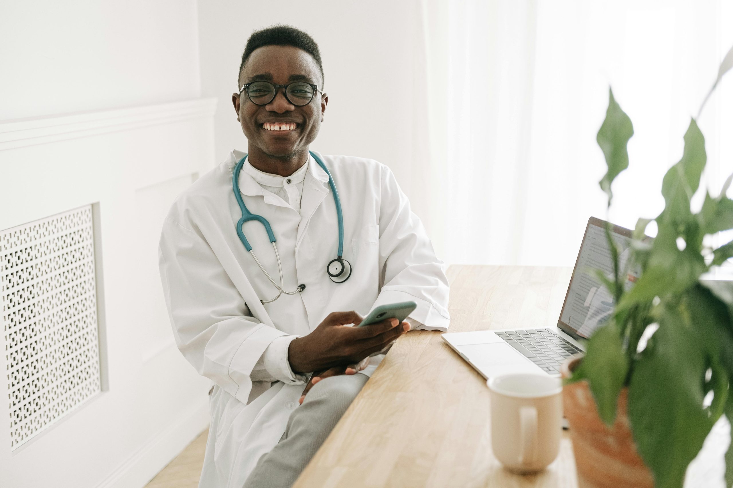 Happy doctor in uniform sitting at a desk using a smartphone in a bright office.