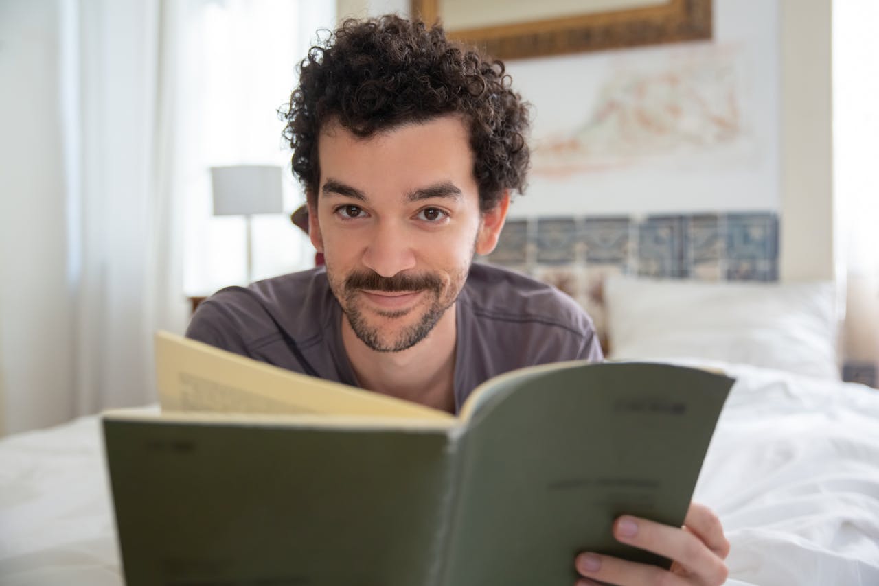 Smiling man with facial hair relaxing and reading a book in a bright, cozy bedroom.